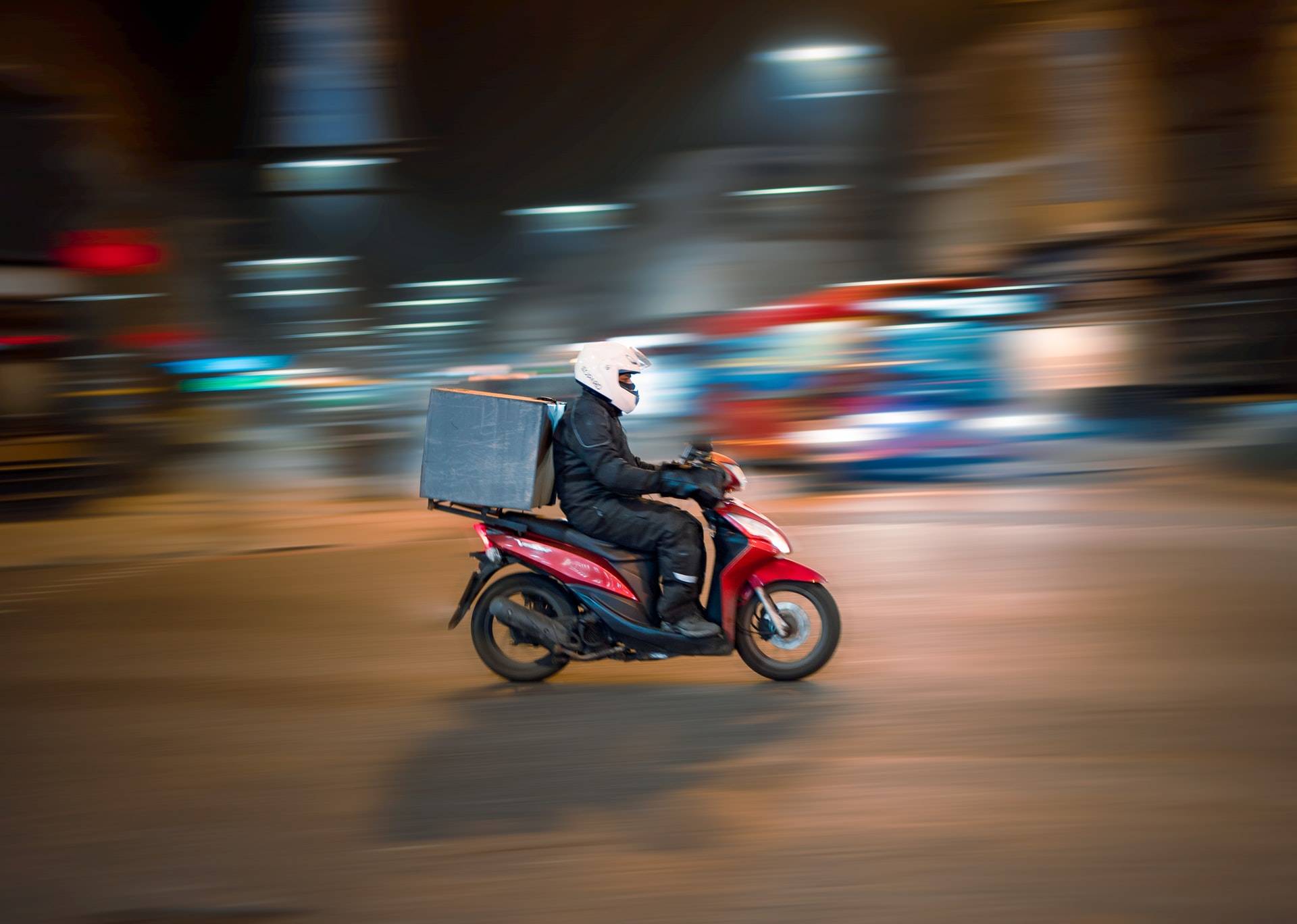 Photo of a man riding a motorised scooter with a luggage box on the back. Scooter and man are in focus while the background is blurred. 